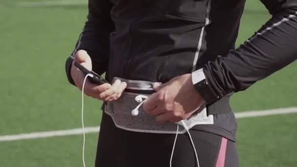 Thème sport et santé. Un jeune homme caucasien utilise un smartphone smarfton pour écouter de la musique sur un casque. Technologie et application mobile playlist sur le tapis roulant de piste de stade de la ville — Video