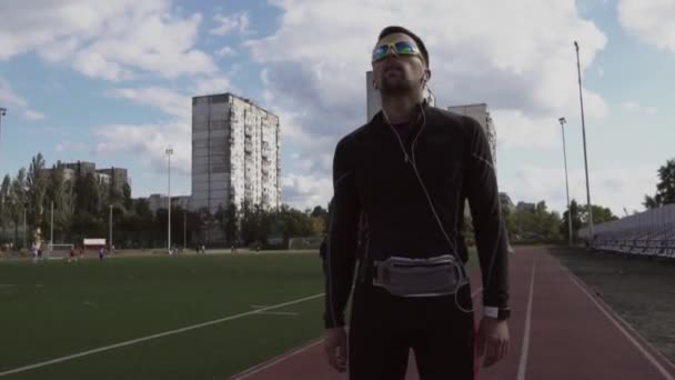 Téma sport és egészség. Egy fiatal kaukázusi férfi portréja, aki fejhallgatóval hallgatja a zenét. Sportlecke a városi stadion futópadján — Stock videók