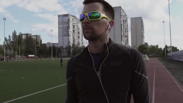 Tema deporte y salud. Retrato de un joven caucásico escuchando música con auriculares. Clase de deportes de entrenamiento en la cinta de correr del estadio de la ciudad — Vídeos de Stock