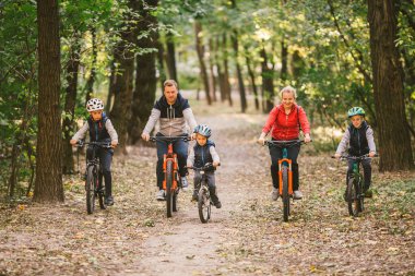Ebeveynler ve çocuklar orman yolunda bisiklet sürüyorlar. Sonbahar parkında bisiklet süren sıcak giysili genç bir aile. Ormanda aile bisikleti. Aile teması: Açık havada spor eğlencesi.