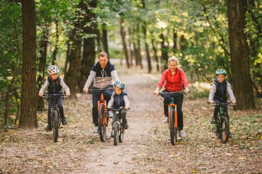 Ebeveynler ve çocuklar orman yolunda bisiklet sürüyorlar. Sonbahar parkında bisiklet süren sıcak giysili bir aile. Ormanda aile bisikleti. Aktif spor, açık hava eğlencesi. 