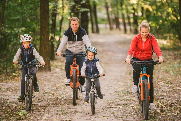 Ebeveynler ve çocuklar orman yolunda bisiklet sürüyorlar. Sonbahar parkında bisiklet süren sıcak giysili bir aile. Ormanda aile bisikleti. Aktif spor, açık hava eğlencesi. 