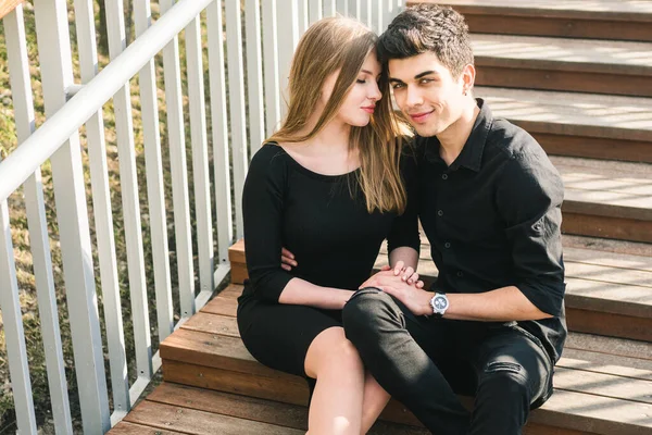 Krásný mladý multiraciální pár, zamilovaný studentský pár, dřevěné schodiště ve městě. Krásná turecká brunetka chlap objímat běloška dívka v černém oblečení. Rande mladí lidé ve slunečném počasí — Stock fotografie