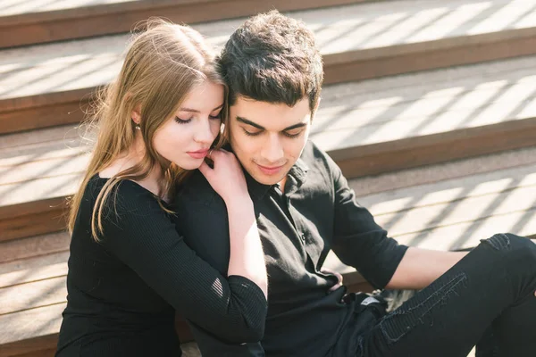 Belo casal multirracial jovem, casal estudante apaixonado, sentar escada de madeira na cidade. Bonito cara morena turco abraçar uma menina caucasiana em roupas pretas. Encontro com jovens em tempo ensolarado — Fotografia de Stock