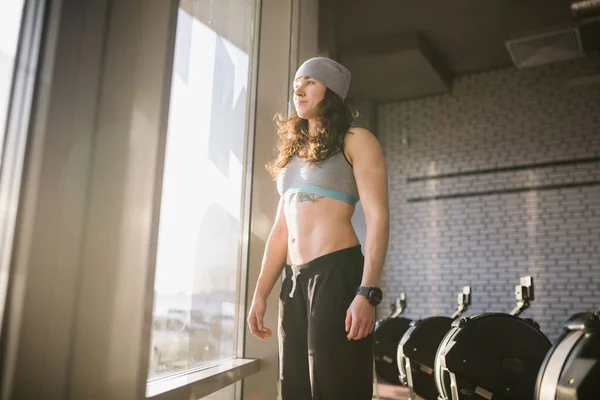 Γυναικείο μοντέλο γυμναστικής στην κορυφή ποζάροντας κοντά σε μεγάλο παράθυρο. γυμναστήριο μοντέλο έχουν διάλειμμα μετά την προπόνηση. Με αυτοπεποίθηση αθλητική γυναίκα με κοιλιακούς που ποζάρουν. Fitness μοντέλο που θέτουν σε εξοπλισμό γυμναστικής θολή φόντο — Φωτογραφία Αρχείου