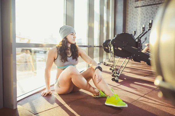 Mujer atlética joven sentada relajándose, descansando entre entrenamientos. Atractiva hembra adulta joven sentada en el piso del gimnasio una ventana doble y posando. slim joven activa chica en ropa deportiva sentarse en el gimnasio — Foto de Stock