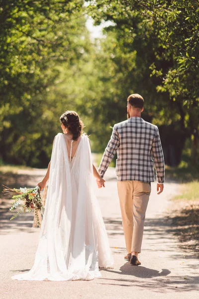 Ευτυχισμένος γαμπρός και νύφη κρατώντας τα χέρια και περπατώντας στην ημέρα του γάμου στον κήπο. Πίσω άποψη του γοητευτικό κομψό νεόνυμφοι κρατώντας τα χέρια, ενώ το περπάτημα στο δάσος πάρκο. Ο δρόμος για τη μελλοντική ζωή. ευτυχισμένες στιγμές γάμου — Φωτογραφία Αρχείου