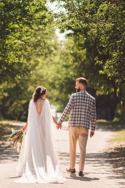 幸せな花嫁と新郎は手を握り、庭の結婚式の日に歩く。公園の森を歩いている間に手を取り合っている魅力的なスタイリッシュな新婚旅行のバックビュー。未来への道幸せな結婚の瞬間 — ストック写真