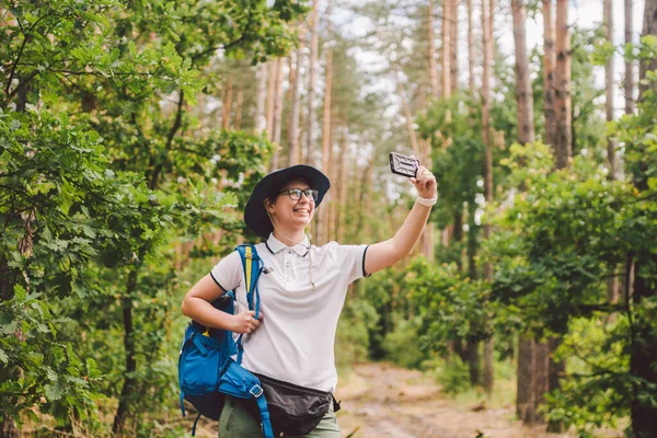 Sorrindo turista feminina detém telefone inteligente, leva selfie contra bela madeira paisagem. Mulher caminhando com mochila tirando foto selfie com smartphone. Viagens e estilo de vida saudável ao ar livre — Fotografia de Stock