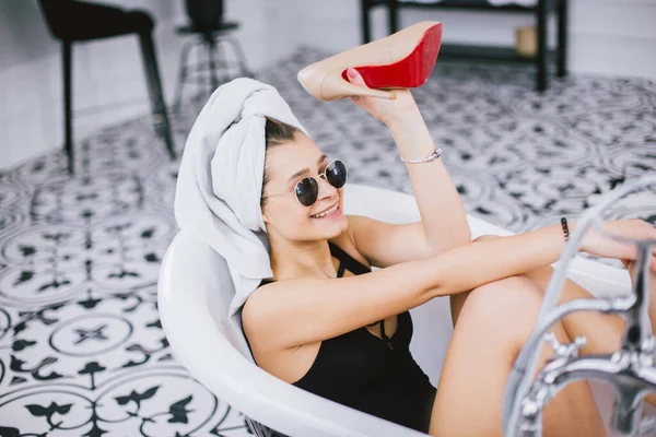 Modeaufnahme vom Einkaufen, junge kaukasische Frau mit Sonnenbrille und Handtuch auf dem Kopf, Stöckelschuhe in der Hand, im Badezimmer am Hotelfenster liegend — Stockfoto