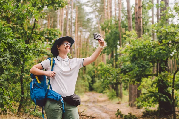 Sorrindo turista feminina detém telefone inteligente, leva selfie contra bela madeira paisagem. Mulher caminhando com mochila tirando foto selfie com smartphone. Viagens e estilo de vida saudável ao ar livre — Fotografia de Stock