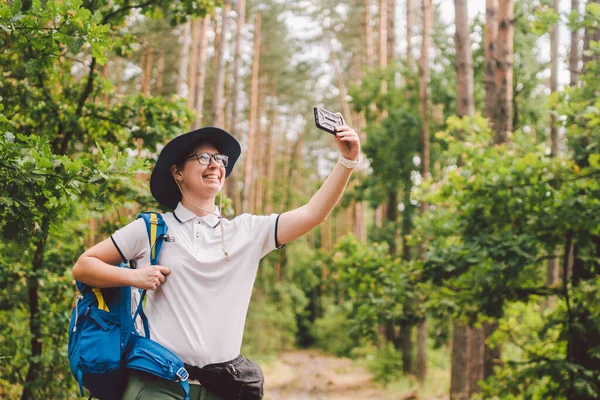 Sorrindo turista feminina detém telefone inteligente, leva selfie contra bela madeira paisagem. Mulher caminhando com mochila tirando foto selfie com smartphone. Viagens e estilo de vida saudável ao ar livre — Fotografia de Stock