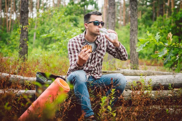 Fast food theme snack on nature. Caucasian man eating a sandwich and drinking water from a palette bottle in the forest. Tourist stopped for lunch in a wooded area