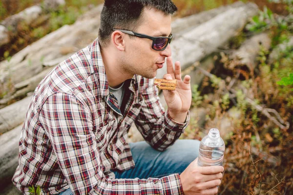 Fast food theme snack on nature. Caucasian man eating a sandwich and drinking water from a palette bottle in the forest. Tourist stopped for lunch in a wooded area