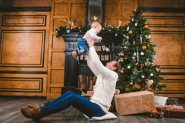 Feliz padre e hijo en casa cerca del árbol de Navidad. Tema Año Nuevo y Navidad. Humor de vacaciones. Concepto de vacaciones de invierno familiar. Padre levanta hijo en brazos sentado en el suelo cerca del árbol de Navidad y regalo — Foto de Stock