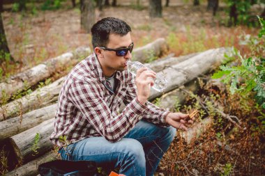 Fast food temalı doğa atıştırması. Kafkasyalı bir adam ormanda sandviç yiyor ve palet şişesinden su içiyor. Turist ağaçlık bir alanda öğle yemeği için durdu.