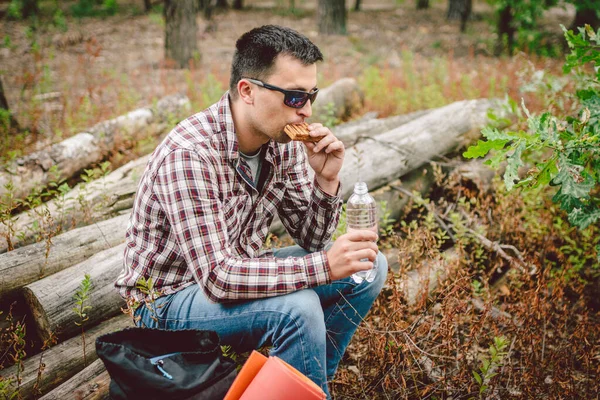 Fast food tematyczne przekąski na łonie natury. Biały mężczyzna je kanapkę i pije wodę z butelki z paletą w lesie. Turysta zatrzymał się na lunch w zalesionej okolicy — Zdjęcie stockowe
