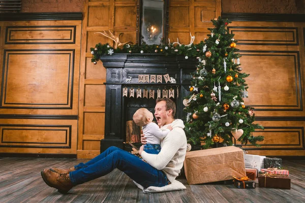 Feliz padre e hijo en casa cerca del árbol de Navidad. Tema Año Nuevo y Navidad. Humor de vacaciones. Concepto de vacaciones de invierno familiar. Padre levanta hijo en brazos sentado en el suelo cerca del árbol de Navidad y regalo — Foto de Stock