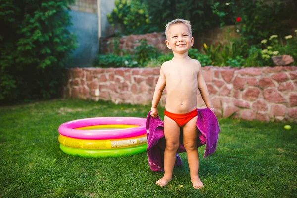 Thème vacances d'été. Un petit garçon caucasien de 3 ans jouant dans la cour d'une maison sur l'herbe près d'une piscine gonflable ronde de couleur enveloppée, essuyée pour être une serviette sèche dans des troncs de natation rouges — Photo