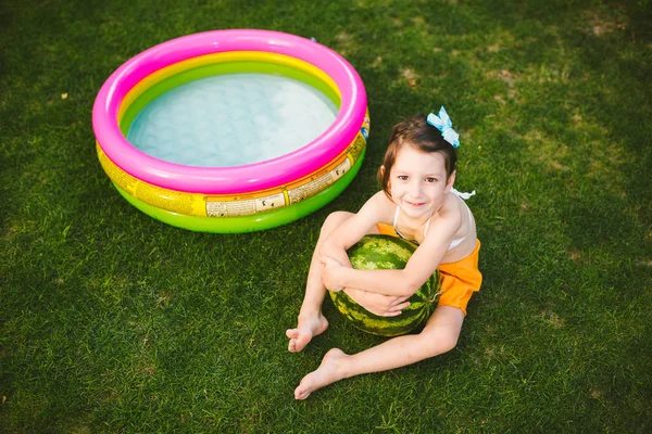 Das Thema gesunde Ernährung von Kindern. kleines Mädchen 4 Jahre alt Kaukasier sitzt auf grünem Gras umarmt große grüne runde Beeren Wassermelone in der Nähe von Kindern aufblasbaren Pool im Hof des Hauses — Stockfoto