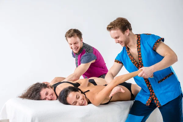 Theme massage and body care. Two young male twin brothers with a beard and uniform make a tandem massage in the hands of two sexy Caucasian women lying on a table on a white isolated background — Stock Photo, Image