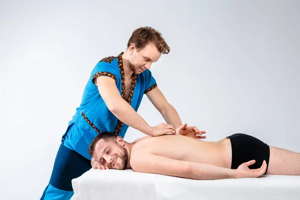 Тематичний масаж і догляд за тілом. Красивий білий чоловік у блакитній формі і борода робить розтягування, лікує, діагностує хлопця з м'язами спини з хорошою фігурою на масажному столі перед білим тлом — стокове фото