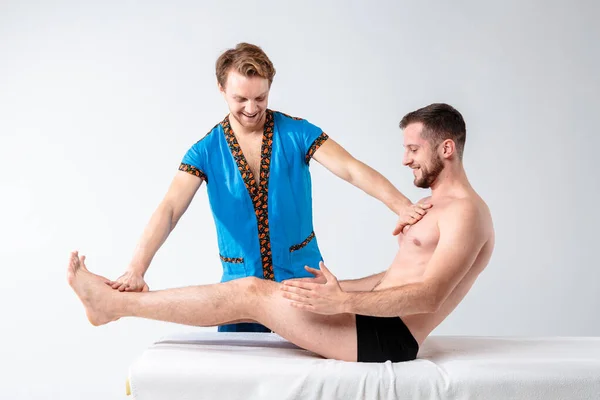 Theme massage and body care. Beautiful caucasian man in blue uniform and beard diagnosing abdominal muscles, stretching guy with good figure on massage table in front of white background — Stock Photo, Image
