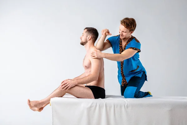 Masaje temático y cuidado corporal. Un guapo médico varón caucásico en uniforme azul y barba diagnosticando músculos de cuello y hombros, estirando chico con buena figura sobre mesa de masaje un fondo blanco — Foto de Stock