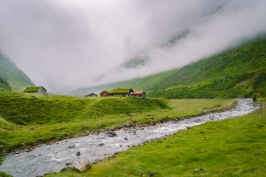 Dağdaki ahşap kulübeler Norveç 'i geçiyor. Tipik İskandinav çim çatı evleri olan Norveç manzarası. Dağ köyü, küçük evleri ve vadide çimleri olan ahşap kulübeleri var.