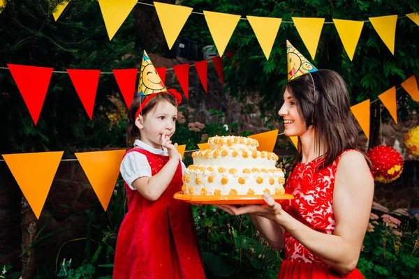 Children's party and cake. A small child of a funny girl is trying to finger a cake which the mother next holds in her hands. Against the backdrop of green trees and festive decor. — Stock Photo, Image