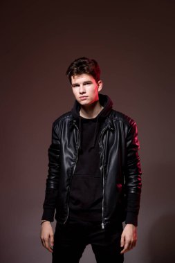 Attractive young guy posing in studio. Young caucasian guy model in black clothes posing in the studio on a dark burgundy background with red backlight. Man in a leather jacket
