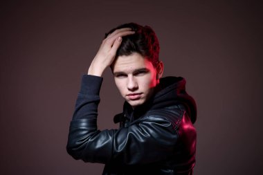 Attractive young guy posing in studio. Young caucasian guy model in black clothes posing in the studio on a dark burgundy background with red backlight. Man in a leather jacket