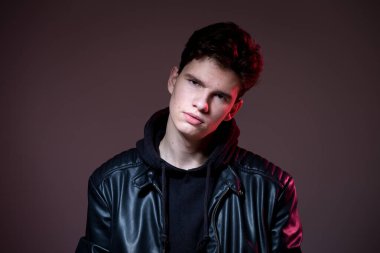 Attractive young guy posing in studio. Young caucasian guy model in black clothes posing in the studio on a dark burgundy background with red backlight. Man in a leather jacket