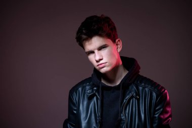 Attractive young guy posing in studio. Young caucasian guy model in black clothes posing in the studio on a dark burgundy background with red backlight. Man in a leather jacket