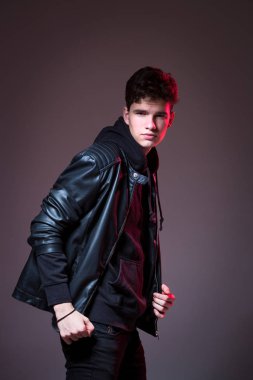 Attractive young guy posing in studio. Young caucasian guy model in black clothes posing in the studio on a dark burgundy background with red backlight. Man in a leather jacket