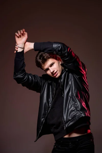 Attractive young guy posing in studio. Young caucasian guy model in black clothes posing in the studio on a dark burgundy background with red backlight. Man in a leather jacket