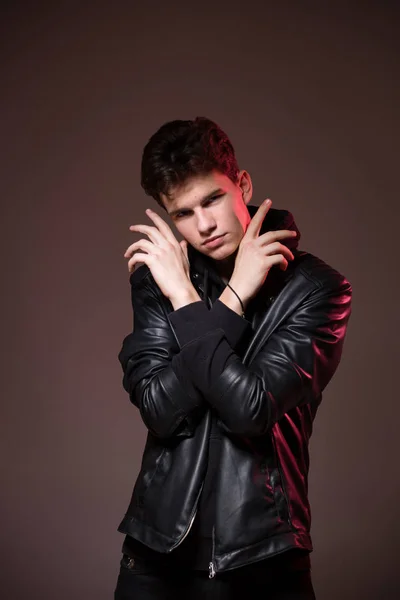 Attractive young guy posing in studio. Young caucasian guy model in black clothes posing in the studio on a dark burgundy background with red backlight. Man in a leather jacket — Stock Photo, Image