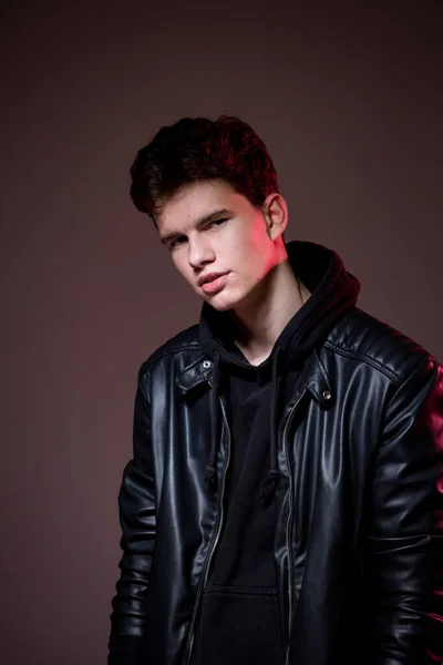 Attractive young guy posing in studio. Young caucasian guy model in black clothes posing in the studio on a dark burgundy background with red backlight. Man in a leather jacket