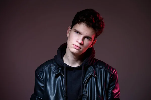 Attractive young guy posing in studio. Young caucasian guy model in black clothes posing in the studio on a dark burgundy background with red backlight. Man in a leather jacket — Stock Photo, Image