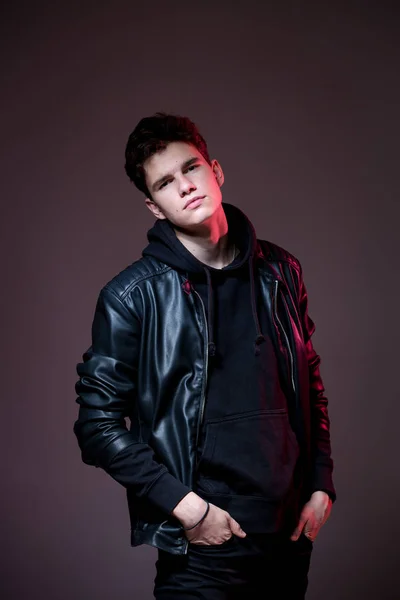 Attractive young guy posing in studio. Young caucasian guy model in black clothes posing in the studio on a dark burgundy background with red backlight. Man in a leather jacket