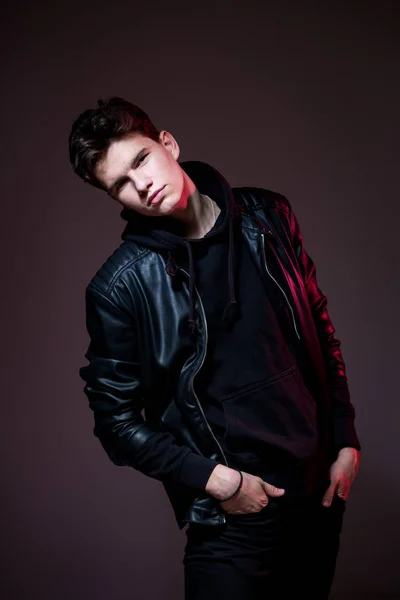 Attractive young guy posing in studio. Young caucasian guy model in black clothes posing in the studio on a dark burgundy background with red backlight. Man in a leather jacket