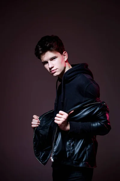 Attractive young guy posing in studio. Young caucasian guy model in black clothes posing in the studio on a dark burgundy background with red backlight. Man in a leather jacket