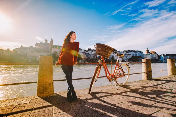 Uma bela jovem está no aterro perto de uma bicicleta da cidade com uma cesta de vermelho na Suíça, a cidade de Basileia. Dia ensolarado na velha Europa — Fotografia de Stock