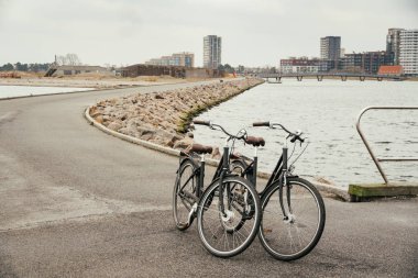 İki özdeş retro siyah şehir bisiklet seti üzerinde park, Baltık Denizi boyunca yol, kış bulutlu havalarda sdan ve şehir Kopenhag Danimarka asfalt yol arka plan ile dolgu