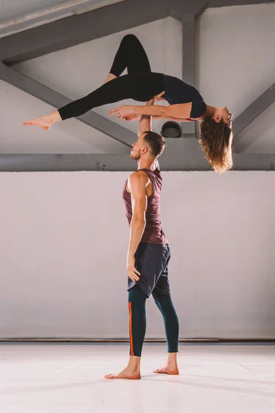 El tema de Acroyoga y Yoga Poses. Un par de dos hombres y una mujer están en la posición de asana. El tipo sostiene a la chica arqueada en el brazo extendido. En el gimnasio con un estudio de contraluz — Foto de Stock