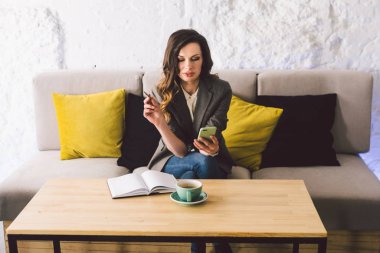 Kahve dükkanında deftere mandıra yazmak, hayatın hatırası olarak. Cep telefonu ve kalemi olan bir kadın kafeteryada bir fincan kahveyle tahta masanın üzerinde. Gülümseyen İş Kadını Not Alıyor