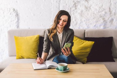 Kahve dükkanında deftere mandıra yazmak, hayatın hatırası olarak. Cep telefonu ve kalemi olan bir kadın kafeteryada bir fincan kahveyle tahta masanın üzerinde. Gülümseyen İş Kadını Not Alıyor