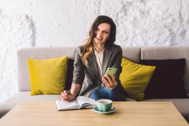 Kahve dükkanında deftere mandıra yazmak, hayatın hatırası olarak. Cep telefonu ve kalemi olan bir kadın kafeteryada bir fincan kahveyle tahta masanın üzerinde. Gülümseyen İş Kadını Not Alıyor