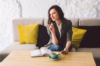 Kahve dükkanında deftere mandıra yazmak, hayatın hatırası olarak. Cep telefonu ve kalemi olan bir kadın kafeteryada bir fincan kahveyle tahta masanın üzerinde. Gülümseyen İş Kadını Not Alıyor