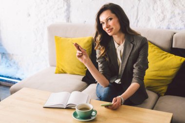 Kahve dükkanında deftere mandıra yazmak, hayatın hatırası olarak. Cep telefonu ve kalemi olan bir kadın kafeteryada bir fincan kahveyle tahta masanın üzerinde. Gülümseyen İş Kadını Not Alıyor